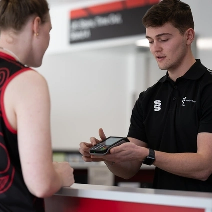 Staff member at a counter presenting a payment terminal to a customer, offering the option to pay in their home currency using Dynamic Currency Conversion